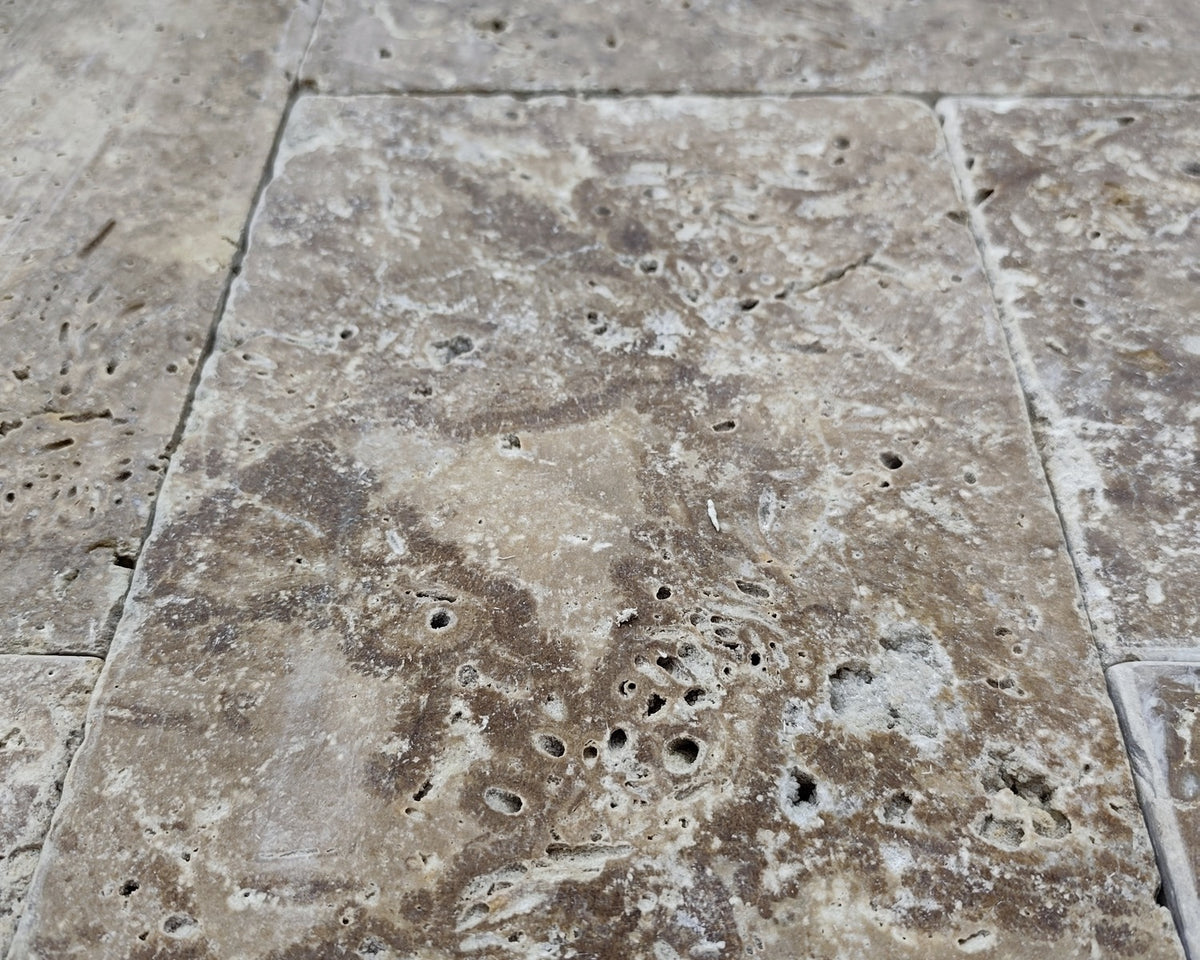 Brown travertine tiles in a Roman pattern with open pores, tumbled edges, and visible tonal variation.