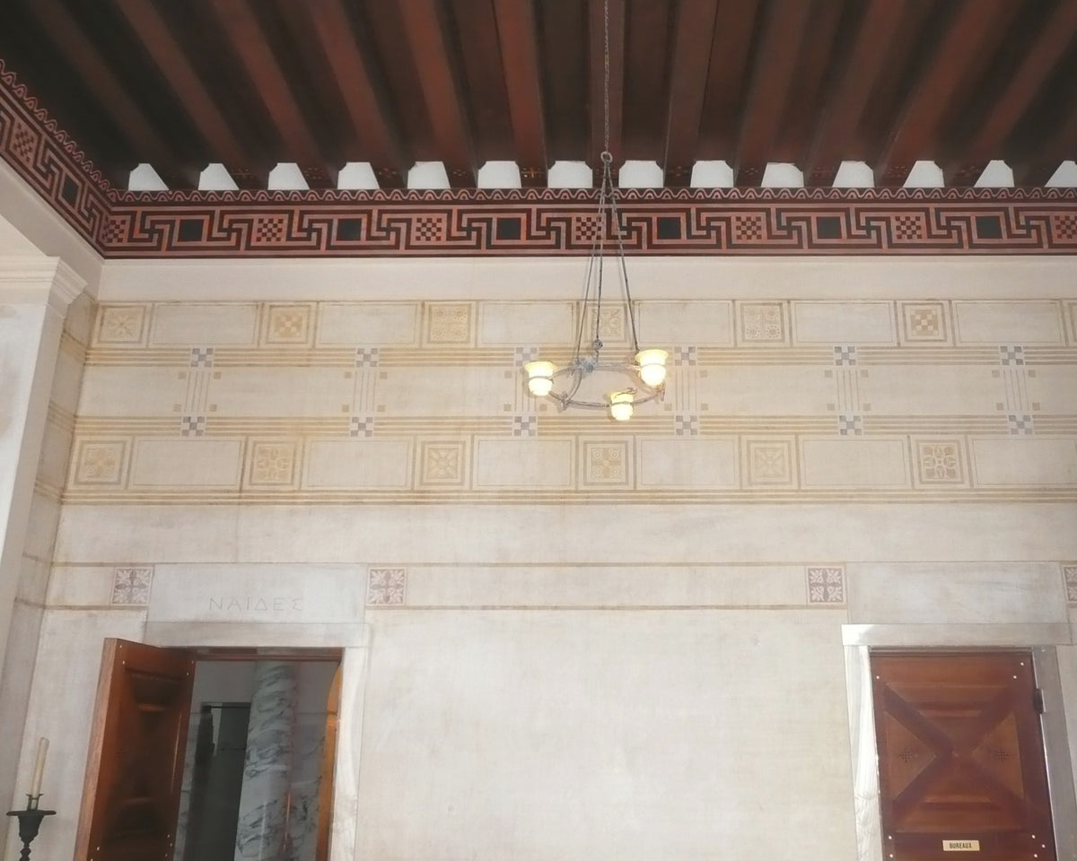 Villa Kerylos interior with a painted Greek-key frieze, patterned walls and a dark beamed ceiling.