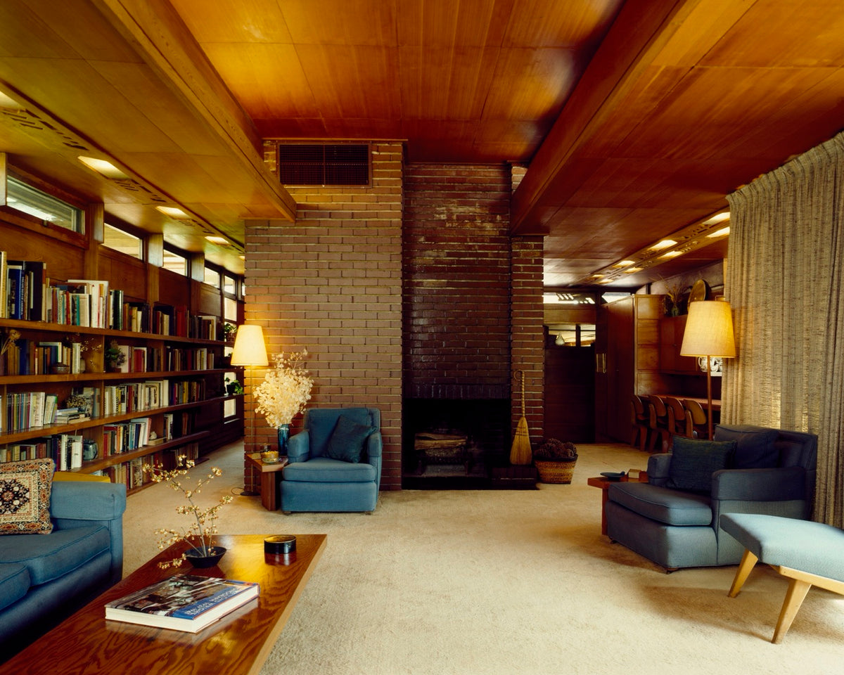 The living room of Frank Lloyd Wright's Rosenbaum House, organized by a central brick hearth, long bookshelves, low seating, and a broad wood ceiling.