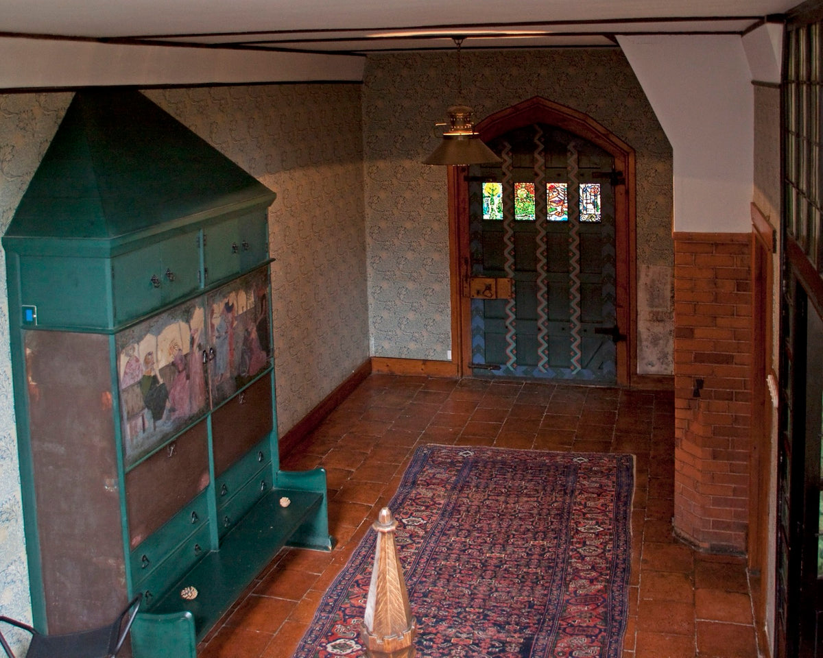 The Red House interior with a full-height green cabinet, patterned rug, brick floor, wallpaper, and a decorated door establishing several levels of scale.